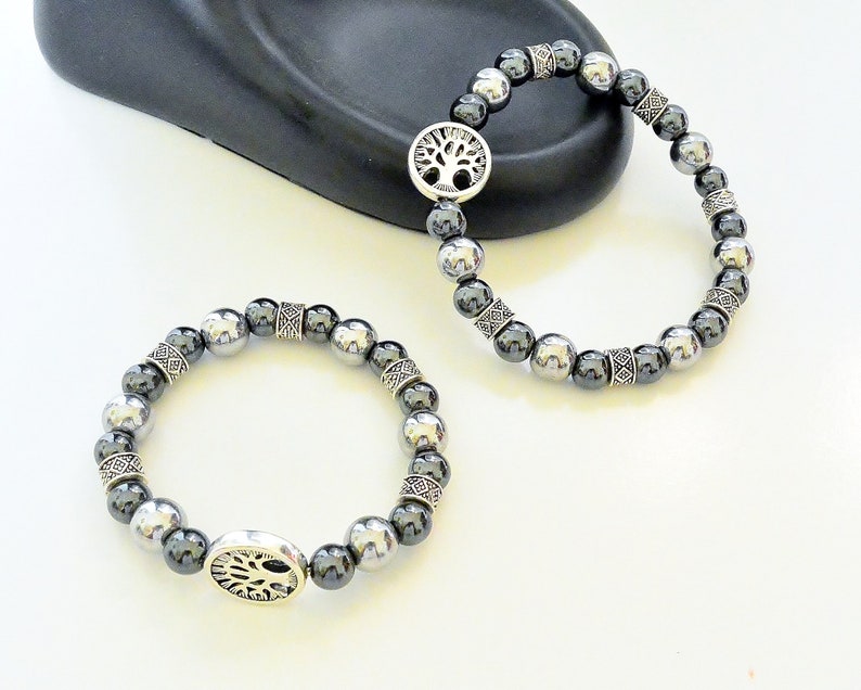 K&ouml;nnte beinhalten: Zwei Armb&auml;nder mit silbernen Perlen und einem silbernen Lebensbaum-Anh&auml;nger. Die Perlen sind schwarz und silber.