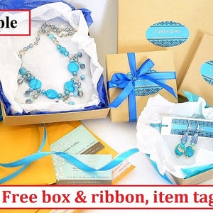 K&ouml;nnte beinhalten: Beispiel f&uuml;r eine blaue und silberne Halskette und Ohrringe, verpackt in einer braunen Schachtel mit einem blauen Band. Die Schachtel ist mit "Cumi's Gems" beschriftet. Das Bild zeigt auch eine Visitenkarte und ein Etikett mit der Aufschrift "Kostenlose Schachtel & Band, Artikel-Etikett".