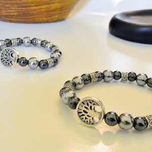 K&ouml;nnte beinhalten: Zwei silberne Armb&auml;nder mit einem Lebensbaum-Anh&auml;nger. Die Armb&auml;nder bestehen aus schwarzen und silbernen Perlen.