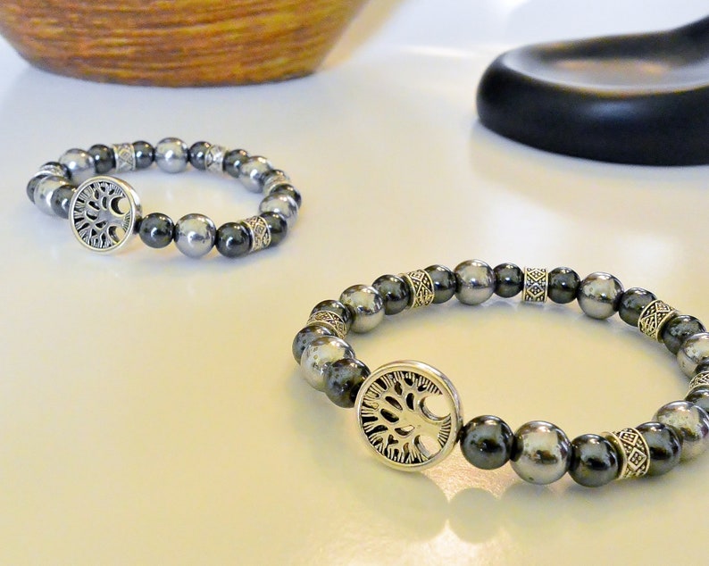 K&ouml;nnte beinhalten: Zwei silberne Armb&auml;nder mit einem Lebensbaum-Anh&auml;nger. Die Armb&auml;nder bestehen aus schwarzen und silbernen Perlen.