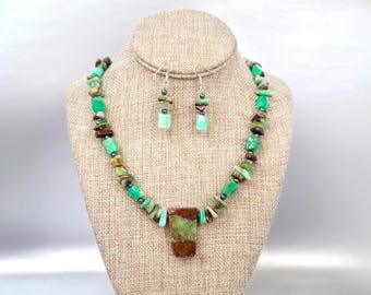 Op de afbeelding: Een ketting en oorbel set met groene en bruine stenen kralen. De ketting heeft een rechthoekige hanger met een groene en bruine steen. De oorbellen hebben een vergelijkbaar ontwerp. De sieraden worden op een mannequin getoond.