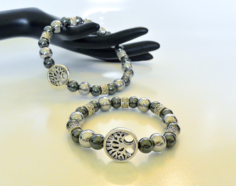 K&ouml;nnte beinhalten: Zwei Armb&auml;nder mit silbernen Perlen und einem silbernen Lebensbaum-Anh&auml;nger. Die Armb&auml;nder befinden sich an einer schwarzen Mannequin-Hand.