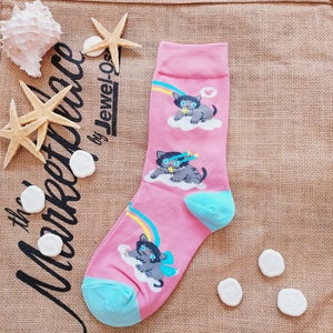 May include: Pink sock with a cartoon cat design. The sock features a light blue heel and toe, with illustrations of gray cats on clouds, rainbows, and stars. The sock is decorated with a bow and a heart.
