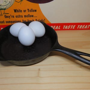 May include: Three white eggs in a black cast iron skillet with a wooden handle. The skillet is on a light wood surface. The text "REAL TASTE TREAT" is visible in the background.