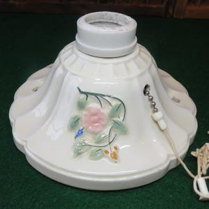 May include: Off-white ceramic ceiling light fixture with a floral design. The fixture has a scalloped edge and a pull chain. The floral design includes a pink flower, green leaves, and small blue and orange accents.