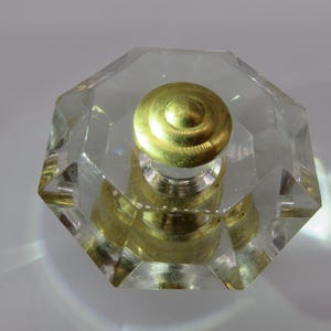 May include: A clear, faceted glass bottle stopper with a gold-colored, spiral-designed top. The stopper has a geometric, octagonal shape and is designed to fit into a bottle. The glass reflects light, creating a sparkling effect.