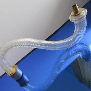 May include: A clear, flexible hose with a ribbed texture. The hose has brass and silver-colored fittings on each end. The hose is curved and appears to be made of a transparent material, reflecting the blue surface it rests upon.