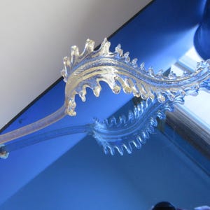 May include: A clear, decorative glass leaf with a stem, mounted on a white base. The leaf has a detailed, textured design with a light gold hue. The object is reflected on a blue surface.