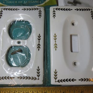 May include: Two rectangular white ceramic wall plates with a gold leaf vine pattern along the borders. One plate is a duplex outlet cover, and the other is a single toggle switch cover. A wooden ruler is visible at the bottom edge of the image.