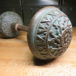 May include: Antique bronze door knob with intricate geometric and floral patterns. The knob has a weathered, aged appearance with a greenish-blue patina. The door knob is made of metal and has a long, slender rod connecting the two decorative ends.