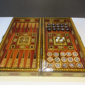May include: A wooden backgammon board featuring intricate marquetry inlay with geometric star patterns. The board is open, displaying a playing surface with alternating dark and light wood triangles, dark and light circular checkers, and four white dice.