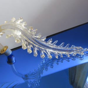 May include: A clear glass decorative object with a leaf-like design. The object has a curved stem with a gold-colored band and a blue base. The object is reflected in a blue surface.