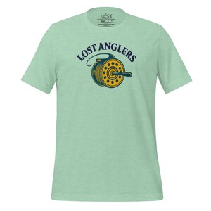 Puede incluir: Camiseta verde claro con las palabras "LOST ANGLERS" sobre un gráfico de carrete de pesca amarillo y azul marino. La camiseta está hecha de un material suave y tiene cuello redondo.