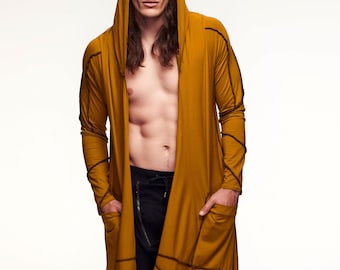 May include: A long, mustard yellow hooded cardigan with black stitching and a slightly asymmetrical hem. The cardigan is open and the model is wearing black trousers and black boots.