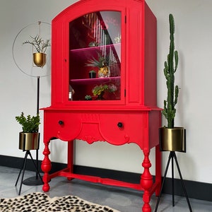 May include: A red vintage china cabinet with glass doors and a single drawer. The cabinet has turned legs and sits on a black stand. The cabinet is decorated with a gold-colored plant and a tiger figurine.