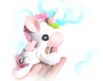 May include: A white felt unicorn with pink and green accents. The unicorn has a pink mane, a green leaf crown, and pink wings. The unicorn is holding a pink heart in its front hoof.