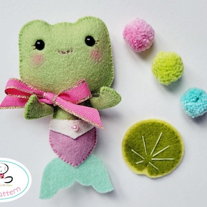 May include: A handmade felt frog mermaid doll with a pink bow, a pink and purple tail, and a pearl. The doll is accompanied by three pom-poms and a felt lily pad. The doll is a PDF pattern.
