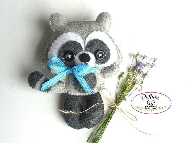 Pu&ograve; includere: Un giocattolo di peluche di procione in feltro grigio con un fiocco a quadri blu e bianco. Il giocattolo ha occhi neri e un naso nero. Il giocattolo tiene un mazzo di fiori viola.
