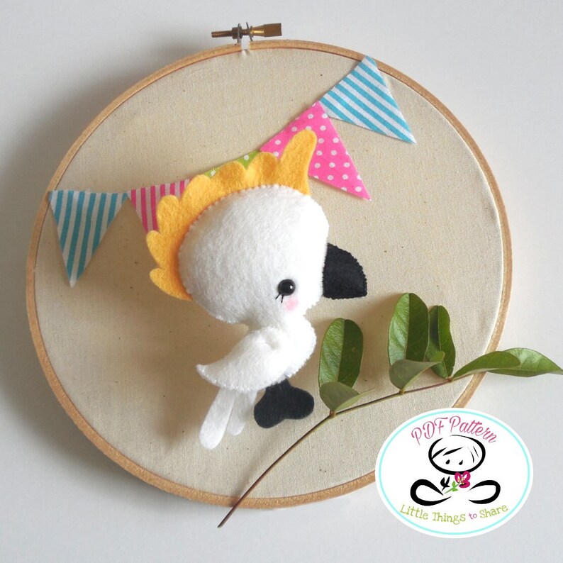 May include: A white felt cockatoo with a yellow crest, sitting on a branch with green leaves. The cockatoo is surrounded by a colorful felt banner with blue, pink, and white stripes. The banner is attached to a wooden embroidery hoop. The image also includes a logo that says "PDF Pattern Little Things to Share".