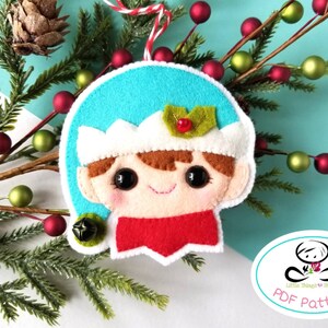 May include: Felt Christmas ornament of an elf with a blue hat, white trim, brown hair, and a red scarf. The ornament has a bell attached to it. The ornament is on a white and blue background with a green sprig of pine.
