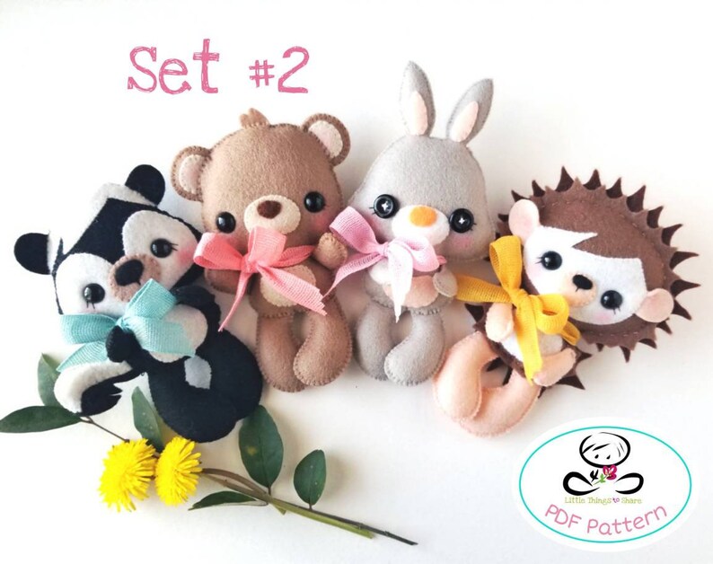 Pu&ograve; includere: Set di quattro peluche di animali in feltro: un orso marrone, un coniglietto grigio, una puzzola bianco e nero e un riccio marrone. Ogni peluche tiene un nastro di un colore diverso. I peluche sono disposti a semicerchio su uno sfondo bianco. Il testo "Set #2" &egrave; scritto in rosa nella parte superiore dell'immagine.