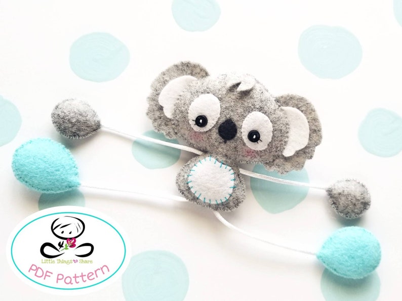 May include: A gray felt koala holding four balloons. Two balloons are gray and two are light blue. The koala has white ears and a white belly with a circle of stitching. The koala is attached to the balloons with white string. The image includes the text "Little Things to Share PDF Pattern".