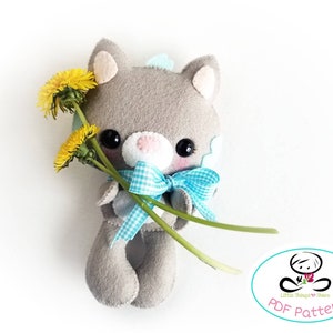May include: A gray felt cat plush toy holding a bouquet of yellow dandelions. The cat has a blue and white checkered bow and black eyes. The image includes the text "PDF Pattern" and a logo with the text "Little Things to Share".