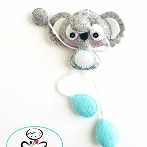 May include: A gray felt koala with white ears and a white felt circle on its belly. The koala is attached to a white string with two turquoise felt teardrop shapes hanging from it. The text "PDF Pattern" is printed in a circle below the koala.