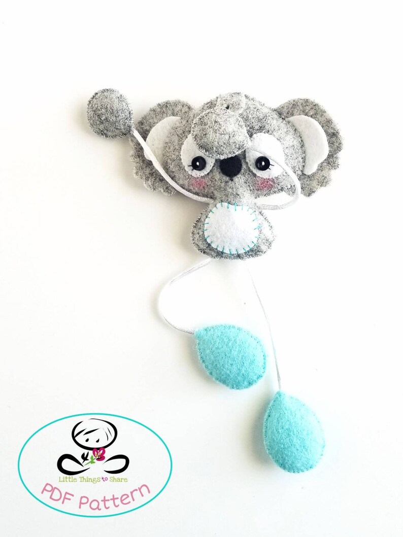May include: A gray felt koala with white ears and a white felt circle on its belly. The koala is attached to a white string with two turquoise felt teardrop shapes hanging from it. The text "PDF Pattern" is printed in a circle below the koala.