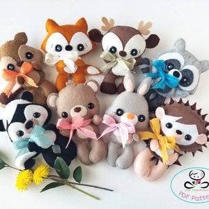 Pu&ograve; includere: Un set di nove peluche di animali in feltro, tra cui una volpe, un cervo, un procione, un orso, un coniglio, un riccio e altri tre animali. I peluche indossano tutti dei fiocchi e sono disposti in cerchio su uno sfondo bianco. Il testo "PDF Pattern" &egrave; visibile nell'angolo in basso a destra dell'immagine.