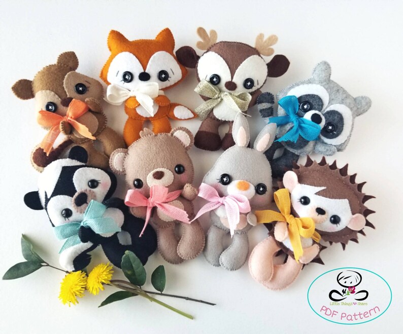 Pu&ograve; includere: Un set di nove peluche di animali in feltro, tra cui una volpe, un cervo, un procione, un orso, un coniglio, un riccio e altri tre animali. I peluche indossano tutti dei fiocchi e sono disposti in cerchio su uno sfondo bianco. Il testo "PDF Pattern" &egrave; visibile nell'angolo in basso a destra dell'immagine.