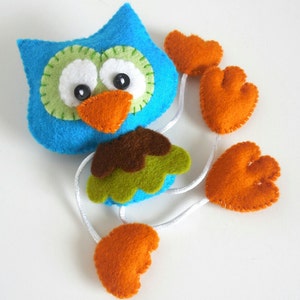 Peut inclure: Un hibou en feutre bleu avec des accents verts et bruns et des pieds en feutre orange. Le hibou a des yeux blancs avec des pupilles noires et un bec orange vif.