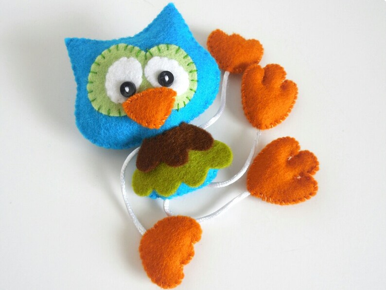 Peut inclure: Un hibou en feutre bleu avec des accents verts et bruns et des pieds en feutre orange. Le hibou a des yeux blancs avec des pupilles noires et un bec orange vif.