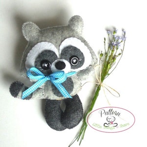 Pu&ograve; includere: Un giocattolo di peluche di procione in feltro grigio con occhi neri e un fiocco a quadri blu e bianco. Il giocattolo tiene un mazzo di fiori viola. Il testo "Pattern Little Things to Share" &egrave; su un'etichetta ovale bianca.