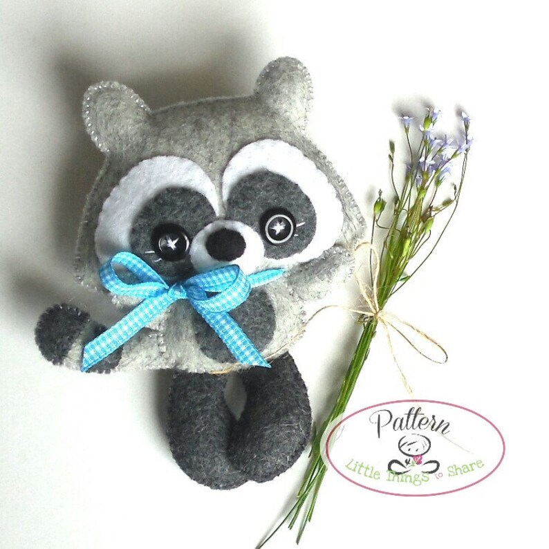 Pu&ograve; includere: Un giocattolo di peluche di procione in feltro grigio con occhi neri e un fiocco a quadri blu e bianco. Il giocattolo tiene un mazzo di fiori viola. Il testo "Pattern Little Things to Share" &egrave; su un'etichetta ovale bianca.