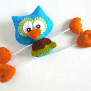Peut inclure: Un hibou en feutre bleu avec des accents verts et bruns et des pieds en feutre orange. Le hibou a de grands yeux blancs et un bec orange vif.