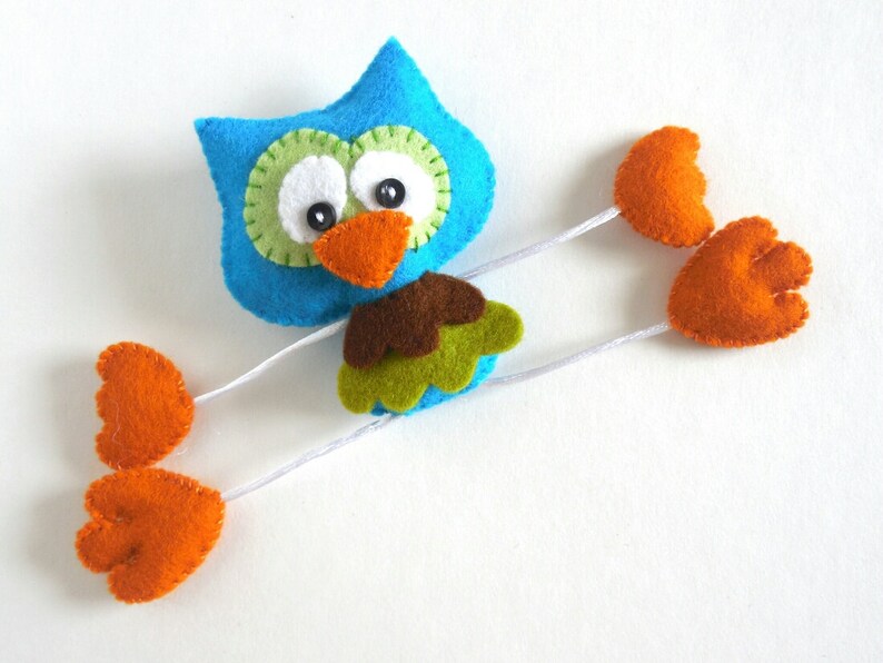 Peut inclure: Un hibou en feutre bleu avec des accents verts et bruns et des pieds en feutre orange. Le hibou a de grands yeux blancs et un bec orange vif.