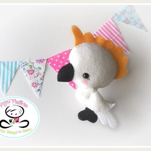 May include: A white felt cockatoo with an orange crest, black eyes, and a black beak. The cockatoo is holding a pink and white polka dot fabric flag. The background is white with a string of fabric flags in various patterns.