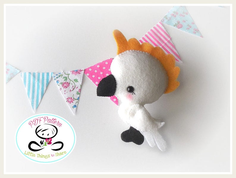 May include: A white felt cockatoo with an orange crest, black eyes, and a black beak. The cockatoo is holding a pink and white polka dot fabric flag. The background is white with a string of fabric flags in various patterns.