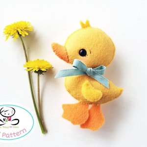 May include: A yellow felt duck with a blue bow, orange feet, and black eyes. The duck is sitting on a white background with two yellow dandelions.