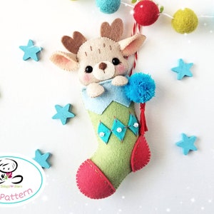 May include: A felt Christmas stocking ornament with a reindeer peeking out of the top. The stocking is green with a blue cuff and red trim. The reindeer is brown with a white face and blue eyes. The stocking has a blue pom-pom on top and a red tassel.