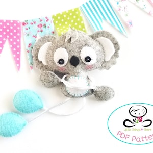 May include: A gray felt koala holding a blue felt balloon. The koala is holding a string with two blue felt balloons attached. The koala is surrounded by a colorful fabric banner.