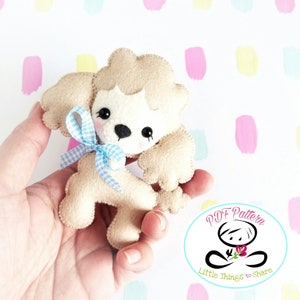 Pode incluir: Um brinquedo de feltro em forma de poodle com um laço azul e branco xadrez. O brinquedo é bege e branco e tem olhos pretos e um nariz preto. O brinquedo está sendo segurado em uma mão. O fundo é uma superfície branca com círculos rosa, amarelos e azuis.