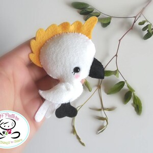 May include: A white felt cockatoo with a yellow crest, black eyes, and a black beak. The cockatoo is holding a small pink flower in its beak. The cockatoo is sitting on a branch with green leaves.
