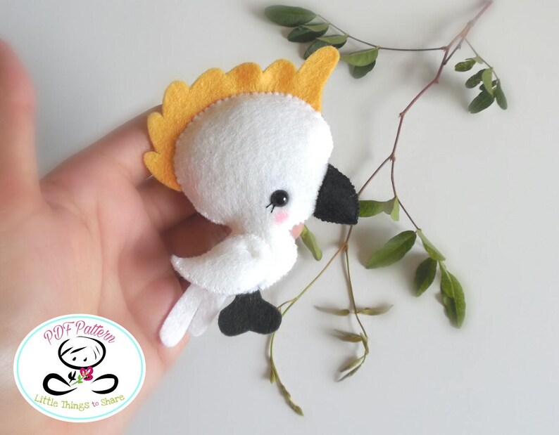 May include: A white felt cockatoo with a yellow crest, black eyes, and a black beak. The cockatoo is holding a small pink flower in its beak. The cockatoo is sitting on a branch with green leaves.