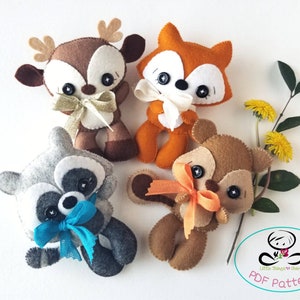 Può includere: Quattro peluche di animali in feltro con fiocchi: un cervo marrone, una volpe arancione, un procione grigio e uno scoiattolo marrone. I peluche sono disposti su uno sfondo bianco con un ramo verde e fiori gialli.