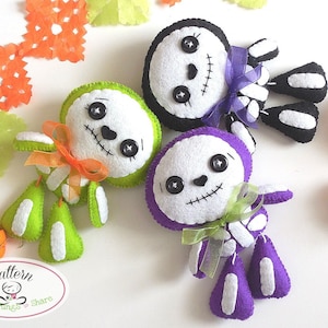 May include: Three felt skeleton ornaments with different colored bodies: green, purple, and black. Each skeleton has a white face with stitched-on details, black button eyes, and a bow.