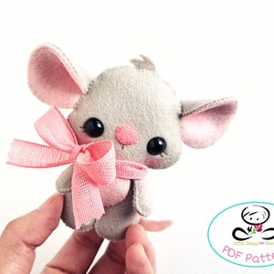 May include: A gray felt mouse with pink ears and a pink bow. The mouse has black eyes and a pink nose. The mouse is being held by a hand.