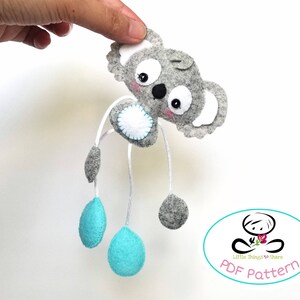 May include: A gray felt koala with blue felt accents and white felt eyes. The koala is hanging from a string and has a white felt circle on its chest. The koala is a PDF pattern for a felt toy.