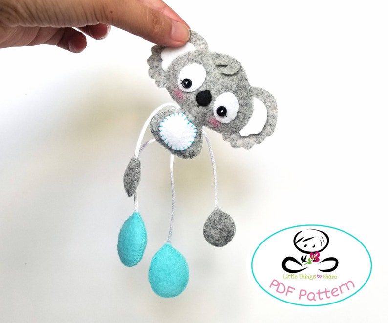 May include: A gray felt koala with blue felt accents and white felt eyes. The koala is hanging from a string and has a white felt circle on its chest. The koala is a PDF pattern for a felt toy.